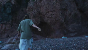 Still image from the video. A human walks towards a seaside cave